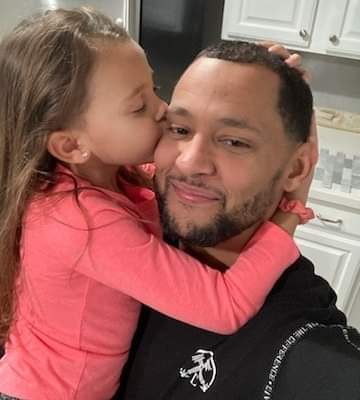 A photo of a smiling Nate with his daughter, and her arms are wrapped around his neck while she kisses him on the cheek.