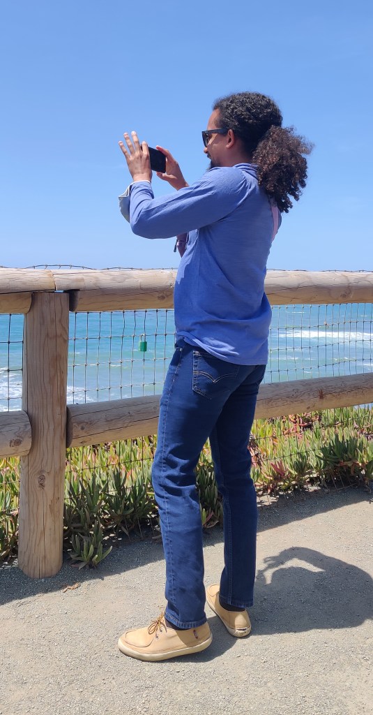 A picture of Ben standing on a lookout point of Moonstone Beach in Cambria, California, filming the view.
